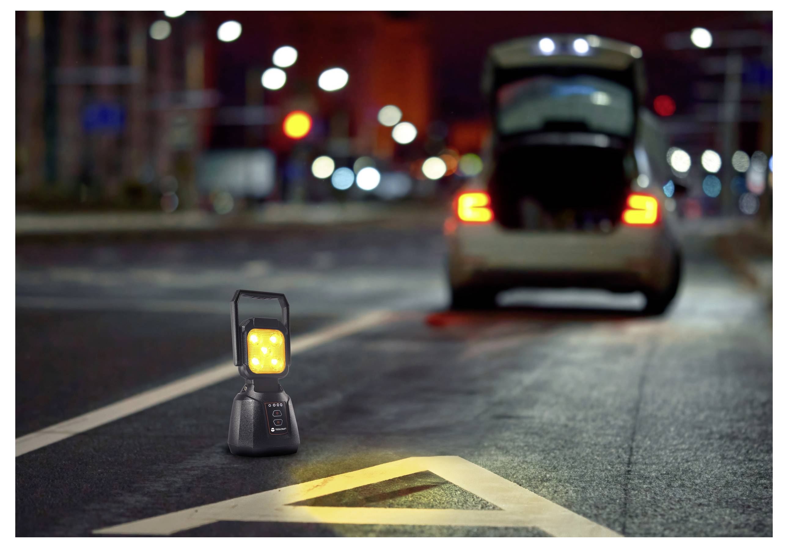 A car with open trunk is parked on a dimly lit street. A portable light with bright LEDs is placed on the road, illuminating the area.