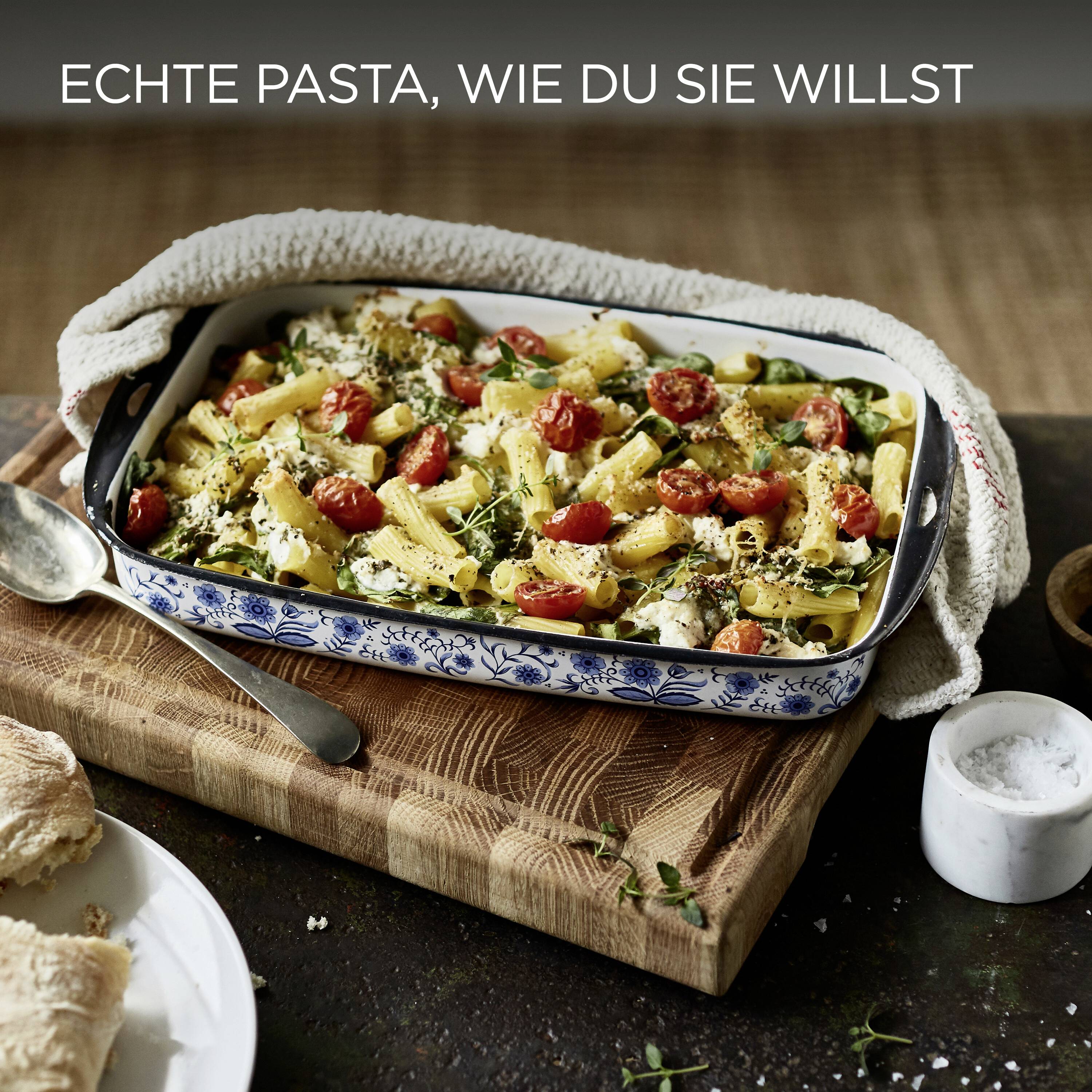 A baked pasta dish with cherry tomatoes, cheese, and herbs in a ceramic dish on a wooden board. Bread pieces and salt are nearby. Text reads 'Echte Pasta, wie du sie willst'.