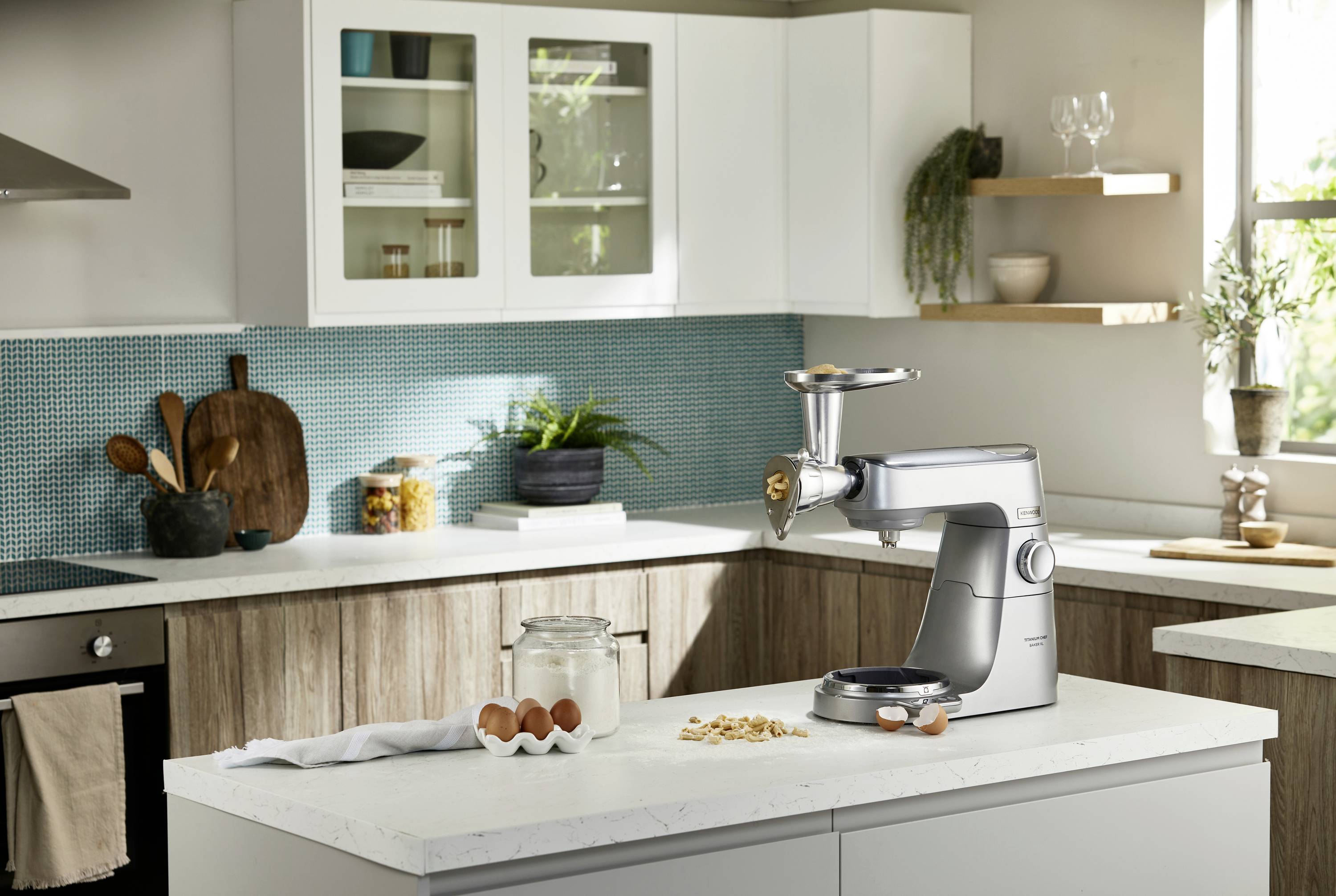 A modern kitchen with white cabinets features a stand mixer with a meat grinder attachment on the counter, surrounded by eggs and flour.