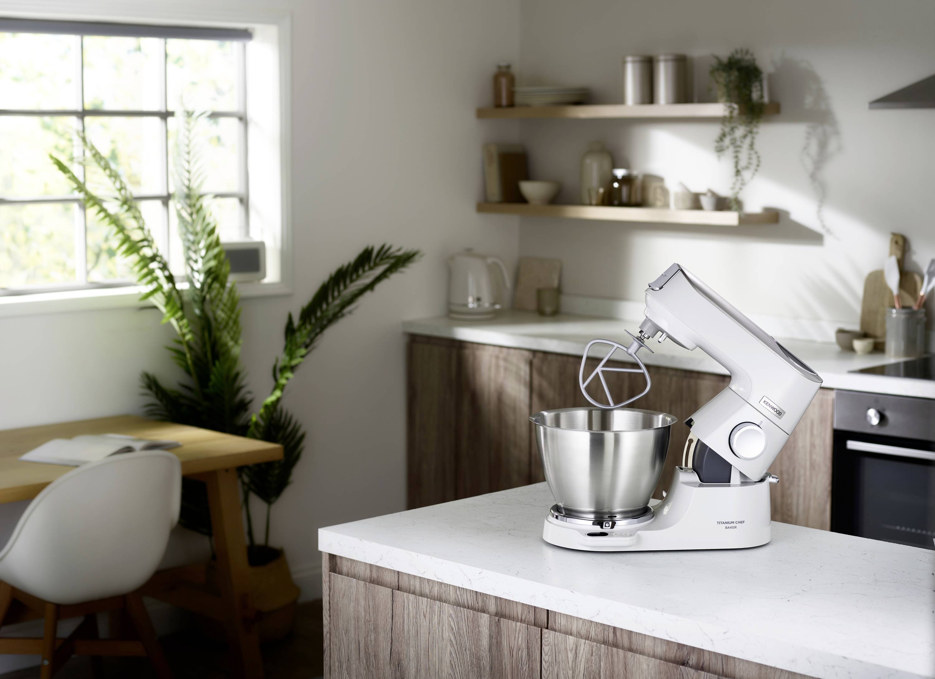 A white stand mixer with a mixing bowl sits on a kitchen counter. The room has wooden cabinets, open shelves, and a window letting in natural light.
