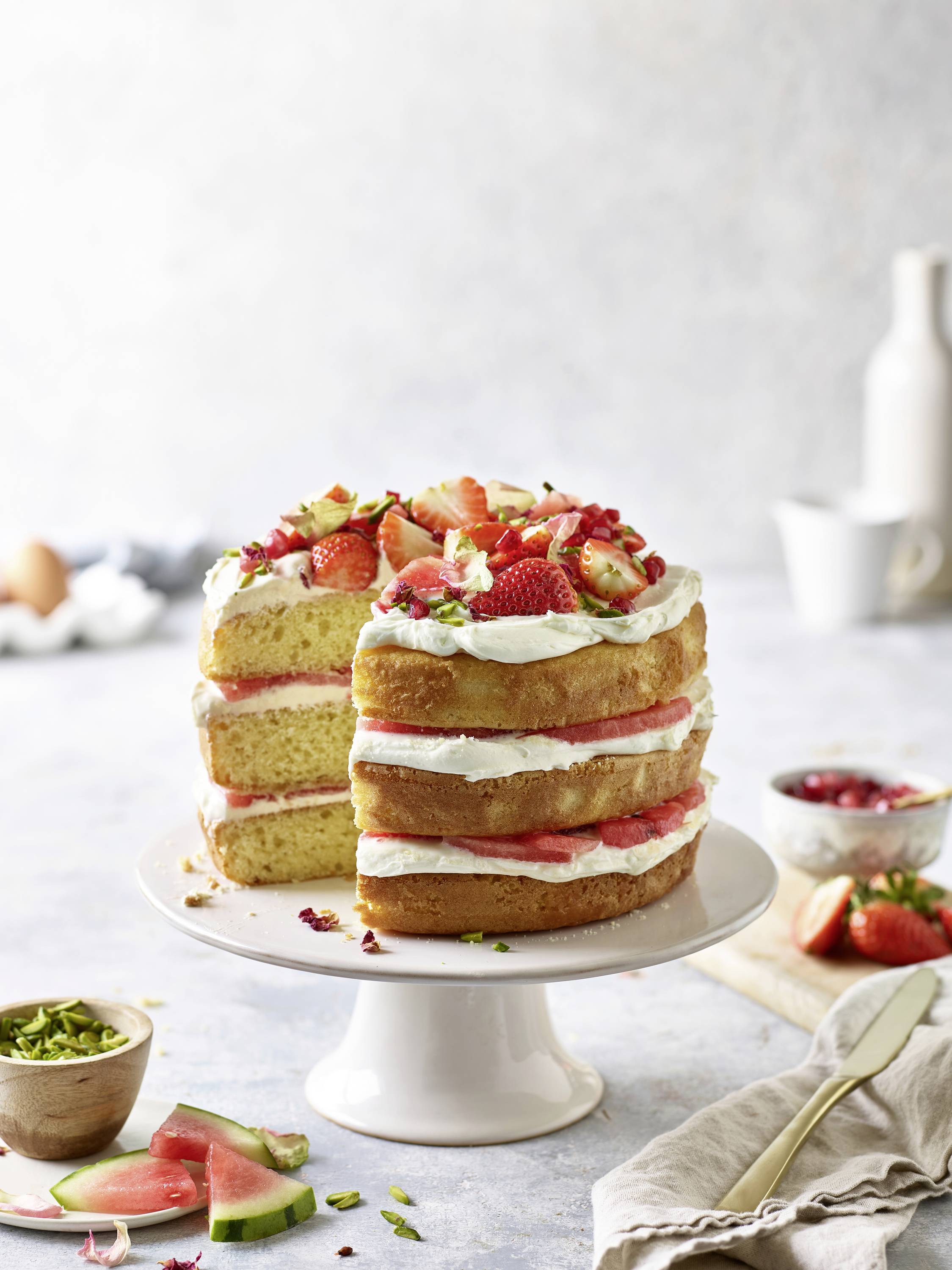 A layered vanilla cake with whipped cream and fresh strawberries, placed on a white cake stand. A fork rests on a napkin nearby.