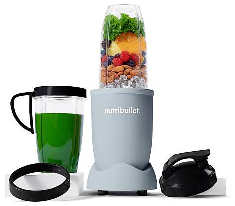 A blender cup filled with almonds, berries, greens, and ice sits on a base next to a green drink in a mug, a lid, and a sealing ring.