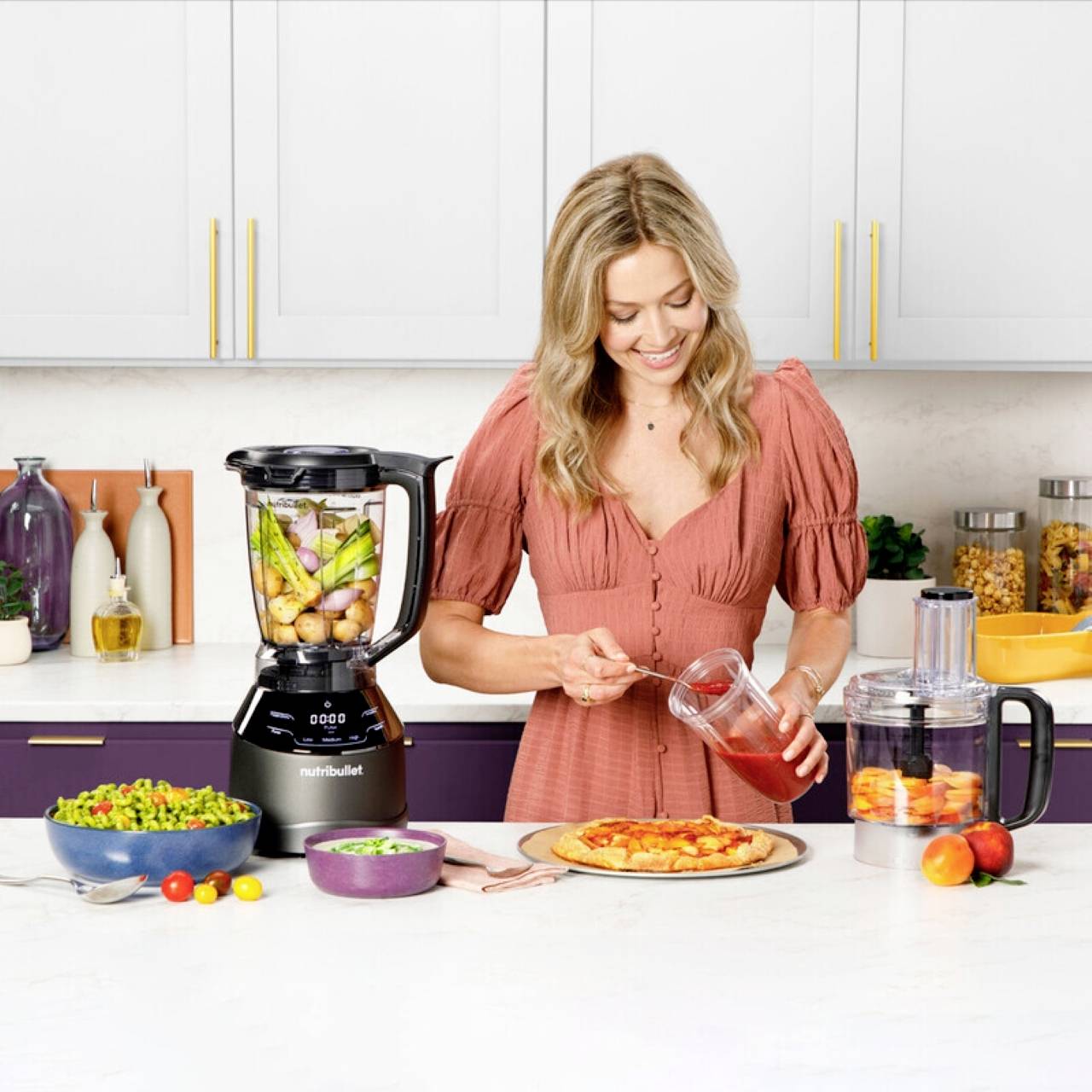 A woman in a kitchen prepares to pour tomato sauce onto a pizza crust. A blender with vegetables and kitchen items are on the counter.