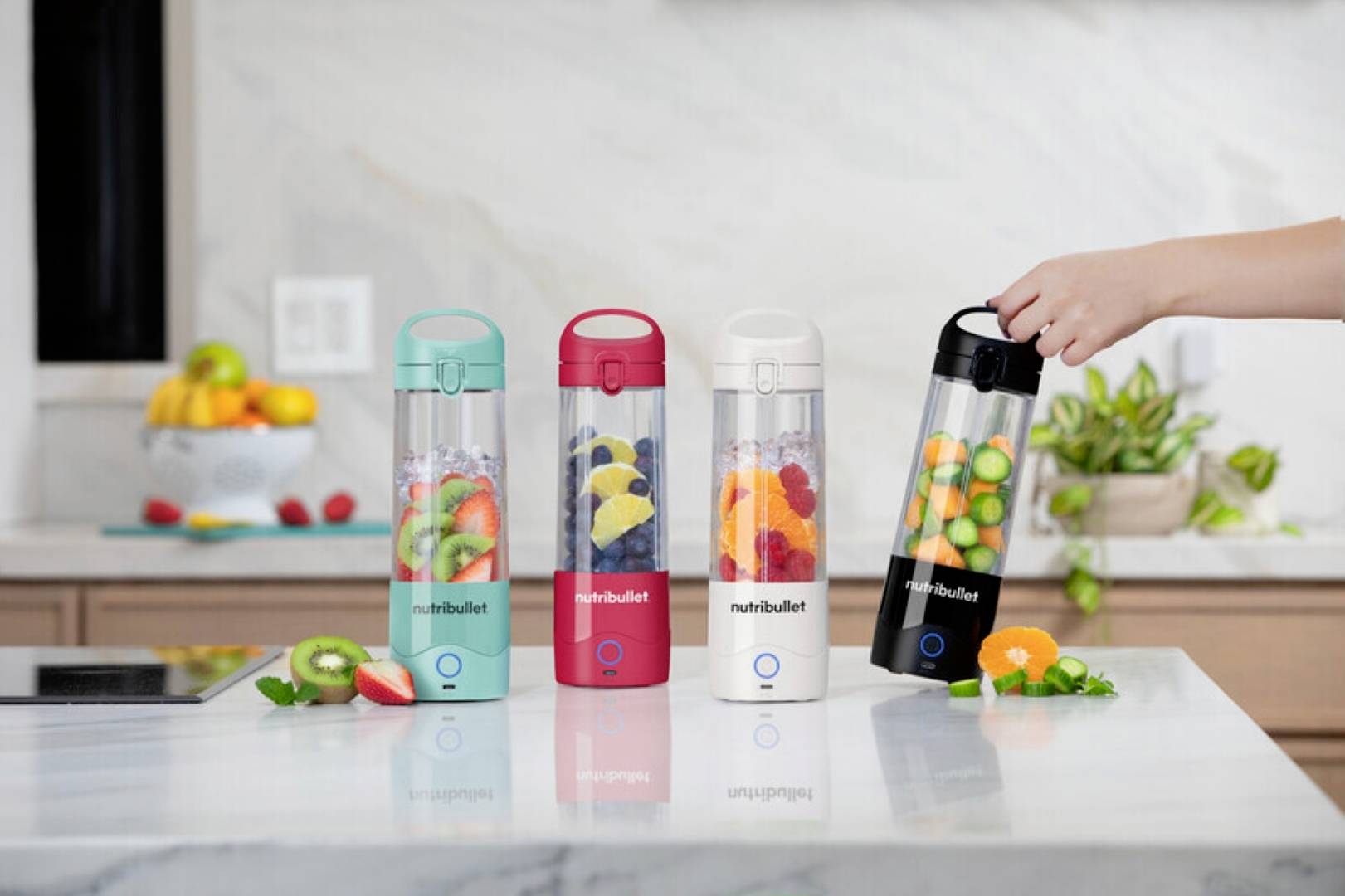 A hand places a portable blender filled with fruits on a kitchen counter beside three other blenders in different colors, all containing fruit.