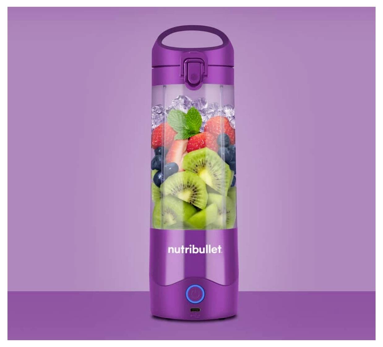 A purple Nutribullet blender bottle filled with ice, kiwi slices, blueberries, strawberries, and mint leaves on a purple background.