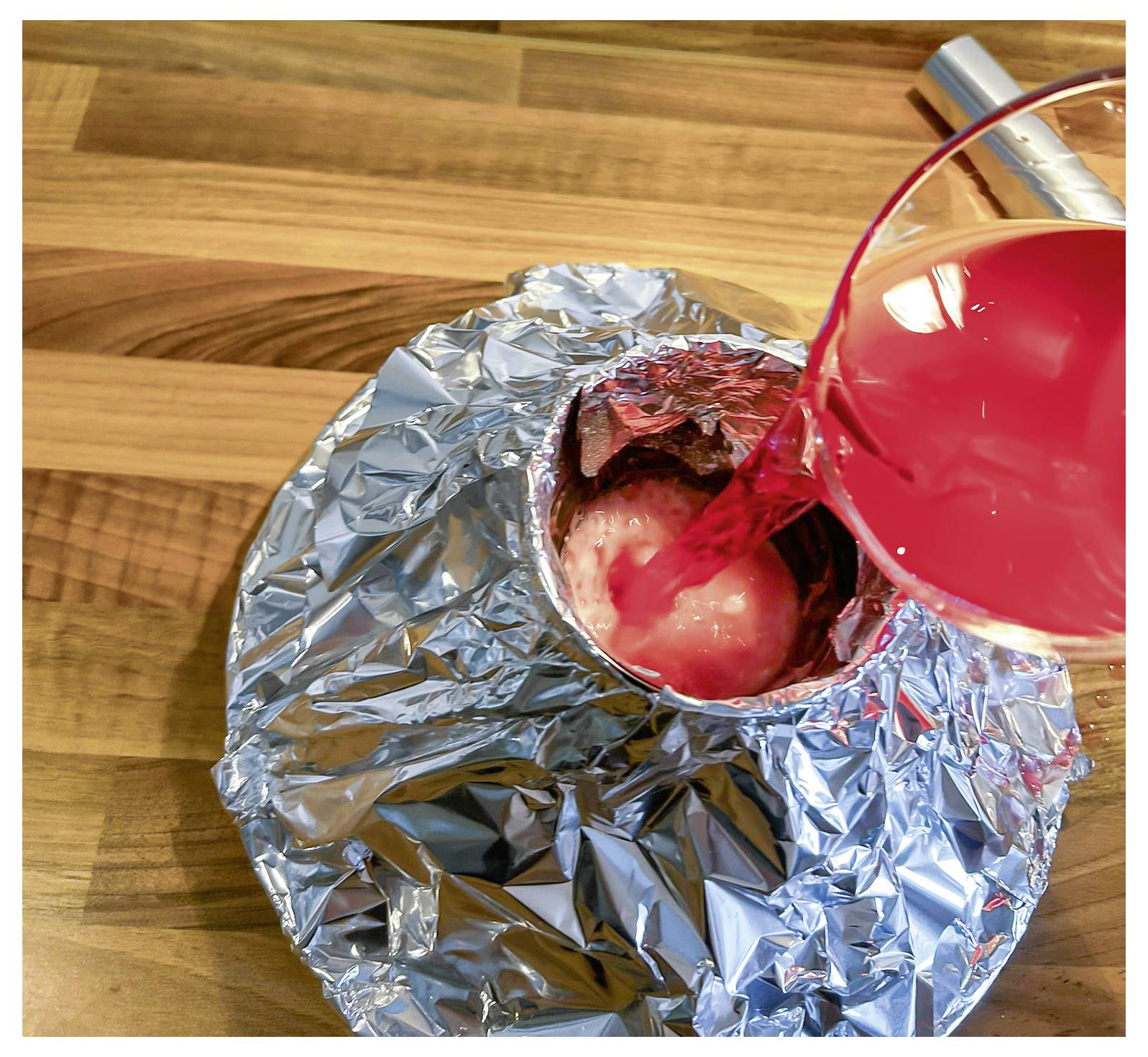 A clear container with red liquid is being poured into a foil-wrapped setup on a wooden surface.
