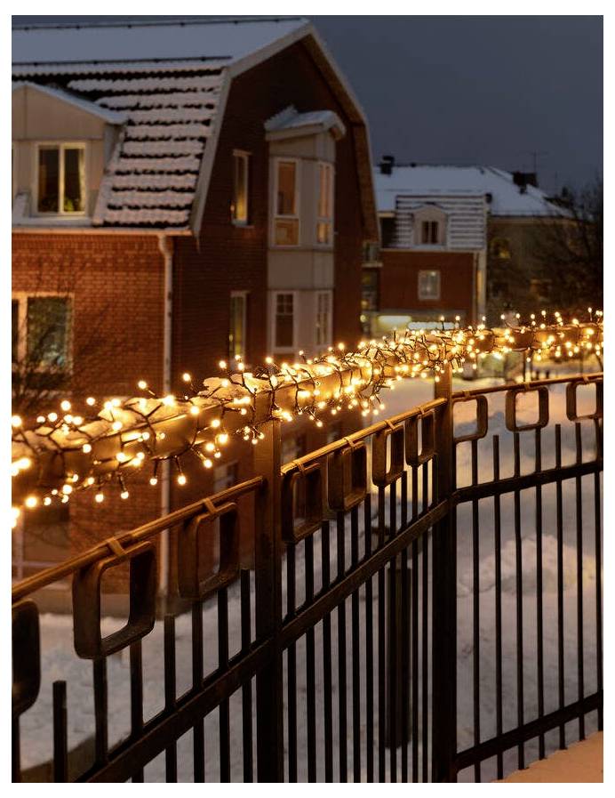 Snow-covered houses and a fence decorated with glowing string lights during the evening, creating a cozy winter atmosphere.