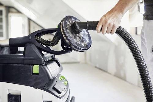 A person using a hand-held sanding tool connected to a vacuum in a bright, partially-renovated room.