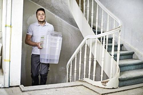 Person carrying a large clear storage bin up a curved staircase in a building under renovation.