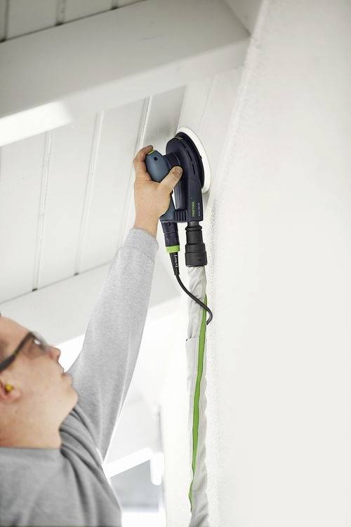 A person using a power sander on a high wall, wearing ear protection and focusing on smoothing the surface.