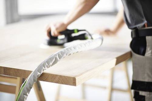 A person using a power sander on a wooden table, smoothing the surface in a workshop setting.