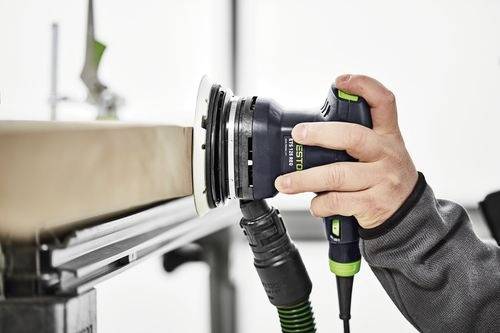 A person using a Festool sander on the edge of a wooden board, smoothing the surface in a workshop setting.