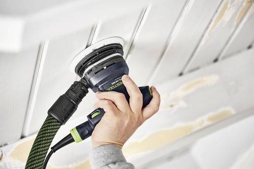 A person is sanding a wooden ceiling with an electric sander, removing old paint. The sander has a green and black handle and is plugged in.