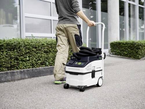 A person pushes a mobile vacuum machine on a sidewalk, next to a building with large windows and trimmed bushes.