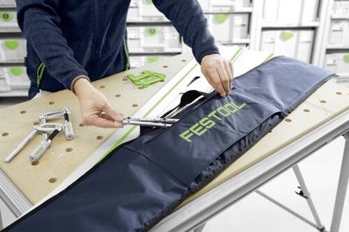 A person assembles tools on a workbench, placing them into a dark blue 'Festool' case. Shelves with containers are visible in the background.