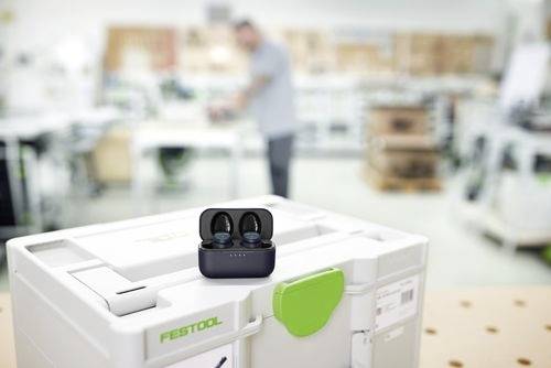 A pair of wireless earbuds placed on a white tool storage box in a workshop setting. A blurred figure is in the background, working.