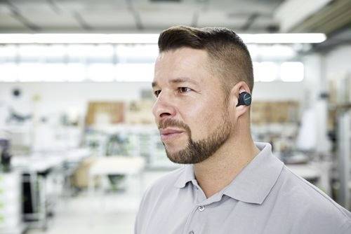 A person wearing a gray collared shirt and a wireless earbud stands in a bright, modern workspace. The background is blurred and industrial.