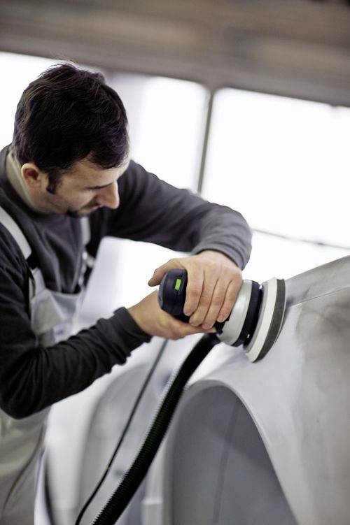 A person wearing protective clothing is using a power sander on a metal surface, likely performing automotive or industrial work.
