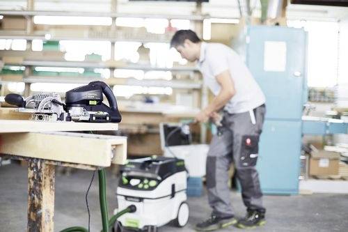 A person in a workshop is operating a vacuum connected to a power tool on a workbench, surrounded by shelves and equipment.
