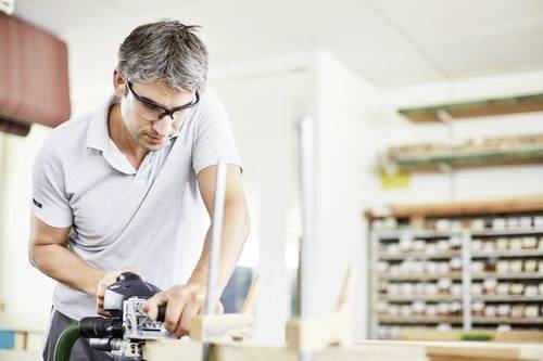 A man wearing safety goggles is using a power tool in a workshop, focusing intently on his work. Shelves with various items are visible in the background.