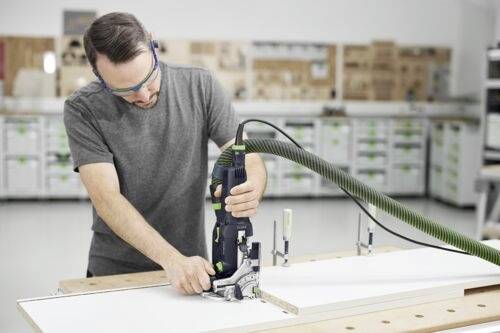 A man wearing safety glasses operates a power tool to cut a piece of wood in a workshop; storage shelves are visible in the background.