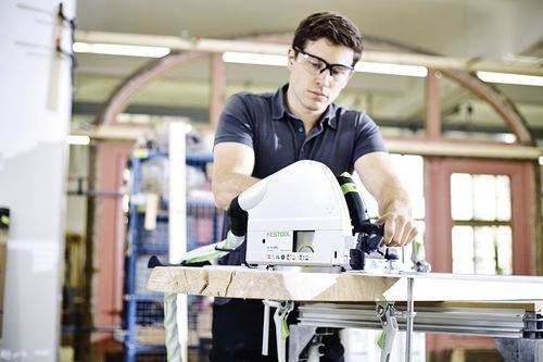 A person wearing safety glasses operates a Festo woodworking saw in a workshop, focusing on precision cutting.
