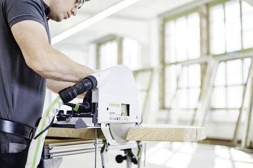 A person operates a circular saw, cutting a wooden board in a bright workshop. Large windows in the background illuminate the space.