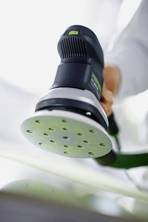 A hand holding a power sander with a green and black handle, viewed from below, displaying its circular sanding pad with holes.