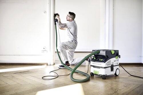 A person kneeling while using a power tool connected to a portable vacuum in a brightly lit room with parquet flooring.
