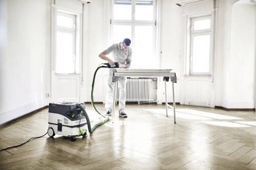 A person sanding a wooden floor in a bright room using a sanding machine attached to a vacuum; windows and white walls are visible.