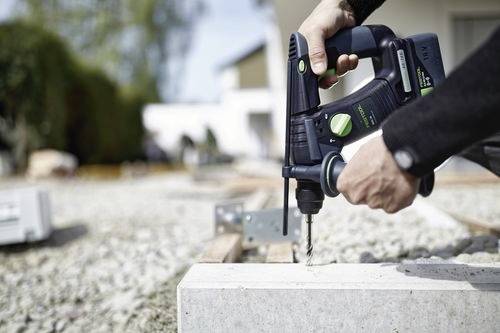 Person using a power drill on a concrete block outdoors, with a blurred background of construction site elements.