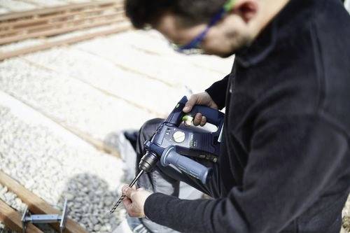 A person in safety glasses uses a power drill on a construction site, focusing on precise drilling into a surface surrounded by gravel.