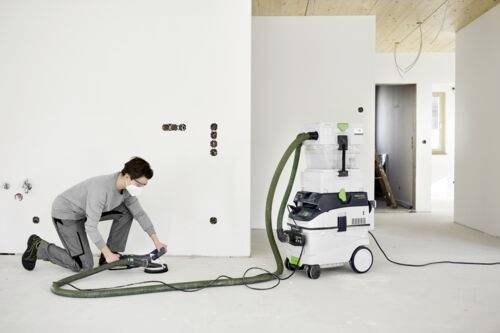 A person wearing a mask uses a handheld sander connected to a vacuum in a partially finished room, suggesting ongoing renovation work.