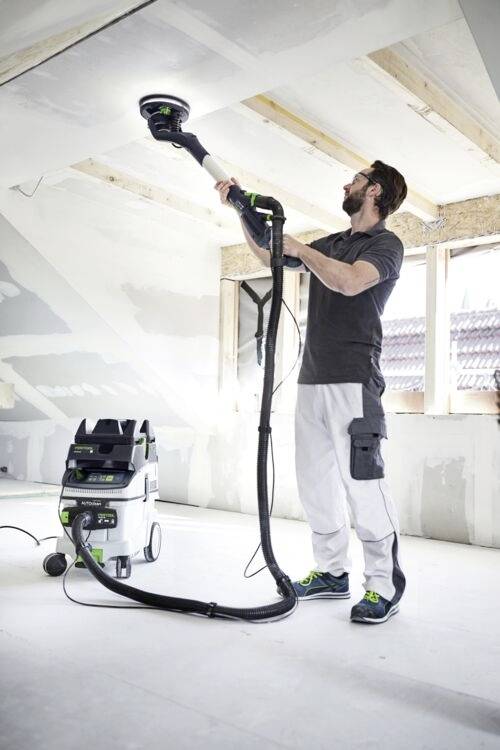 A person in a work outfit uses a power sander on a ceiling in a partially renovated room. Nearby, a vacuum cleaner collects the dust.
