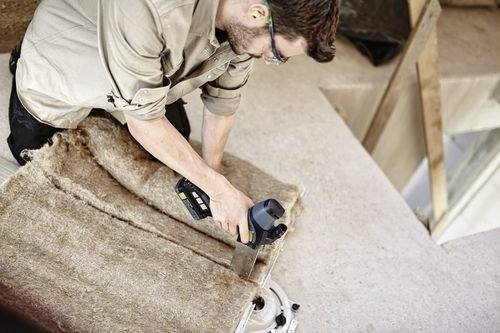 A man in safety glasses and ear protection uses a power tool to cut a roll of insulation material on a workbench in a workshop.