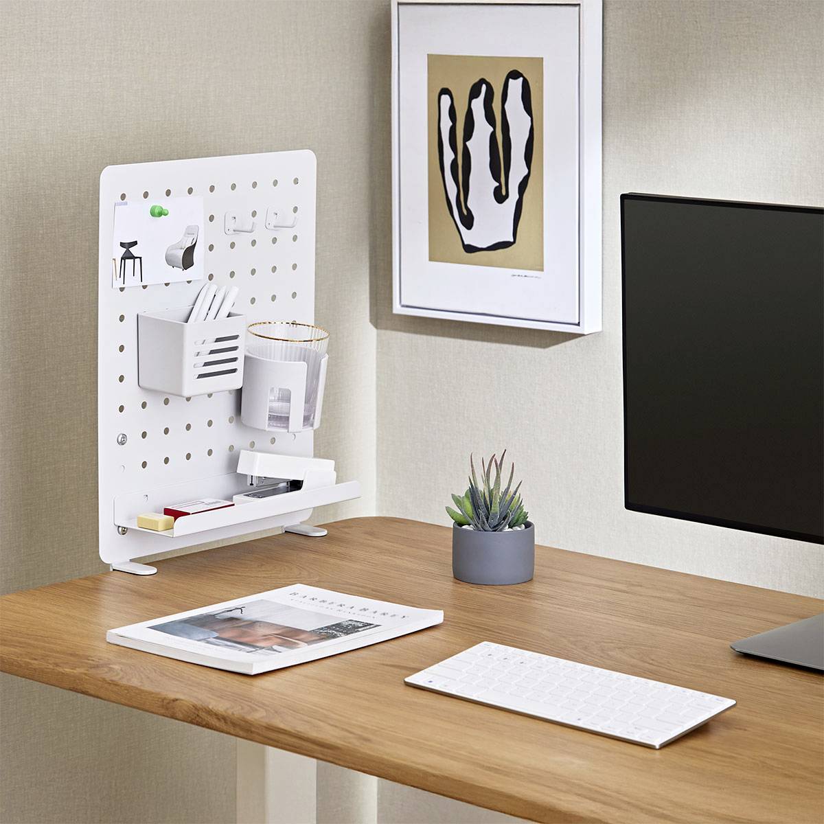 Modern office desk with pegboard organizer, plant, and framed abstract art on wall; minimalist workspace setup with keyboard.