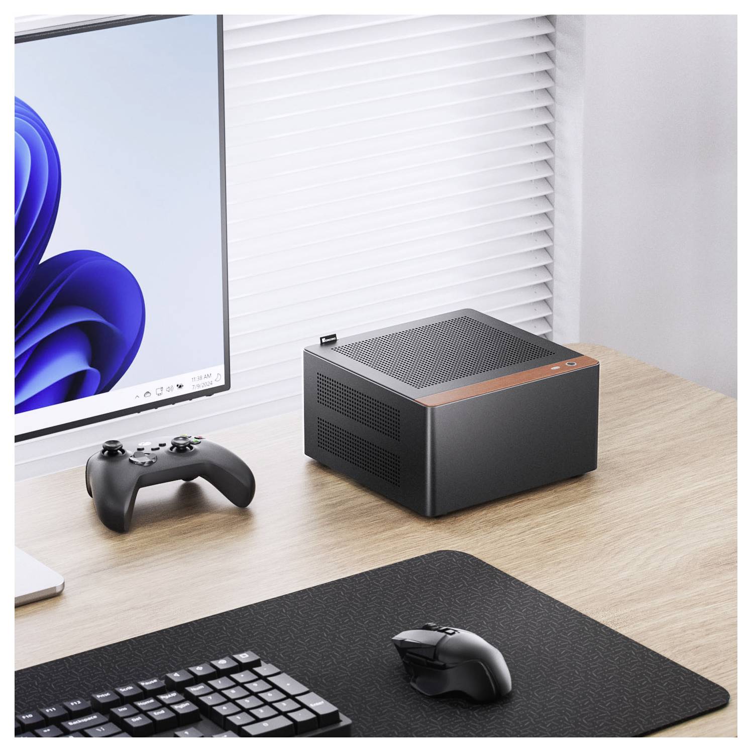 A compact, modern desktop computer on a wooden desk beside a gaming controller and keyboard. A monitor displays a blue swirl wallpaper.