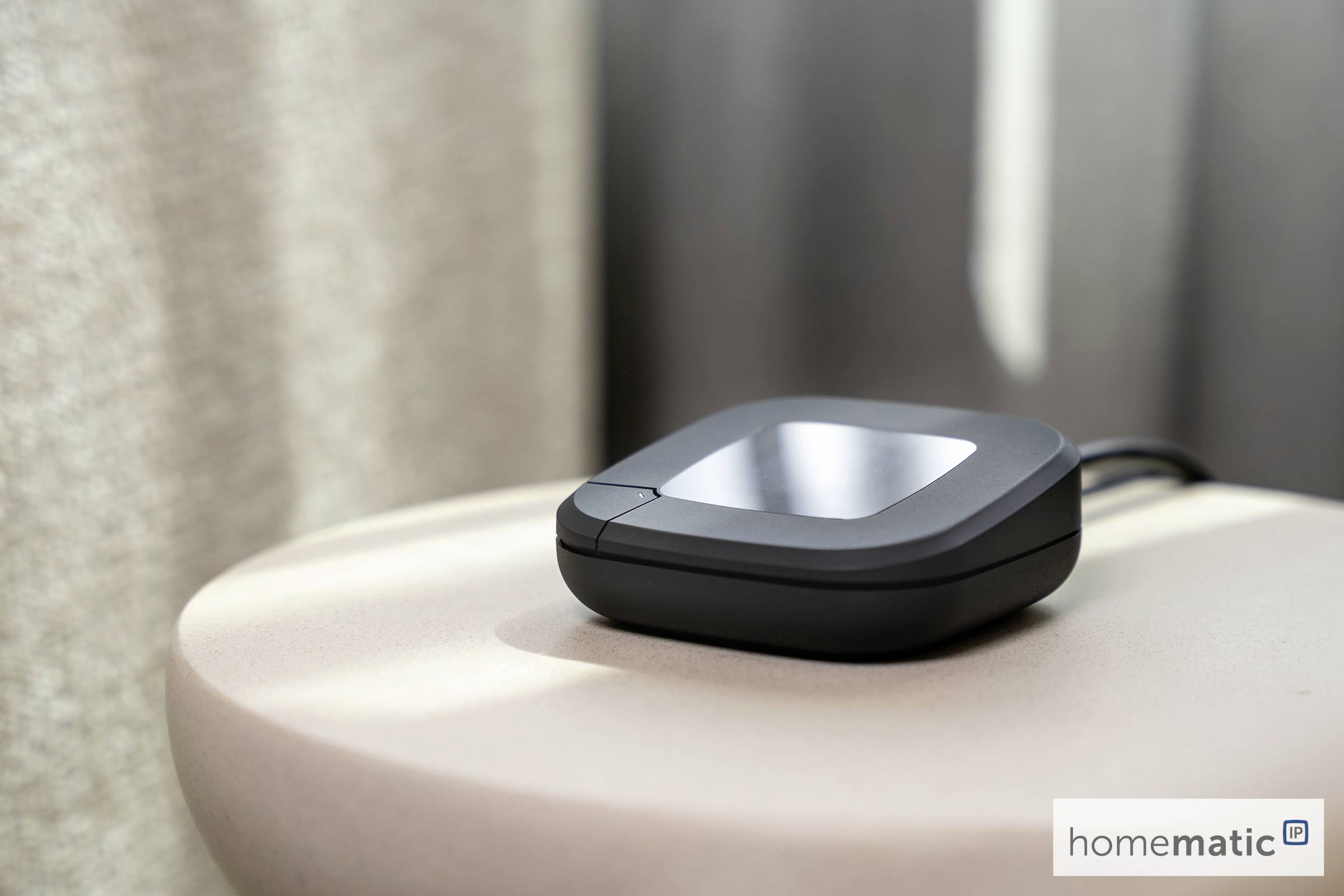 A small, black, square electronic device sits on a round, light-colored table, with a cable attached. A label reads 'homematic.'