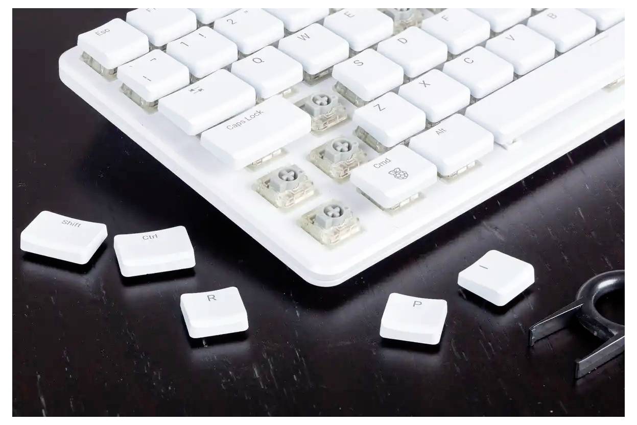 A close-up of a white keyboard with several keys removed, exposing the switches underneath. A keycap puller and loose keys are nearby.
