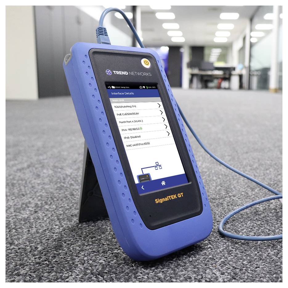 A blue network tester device with a display screen showing interface details, standing on a carpeted floor in an office setting.