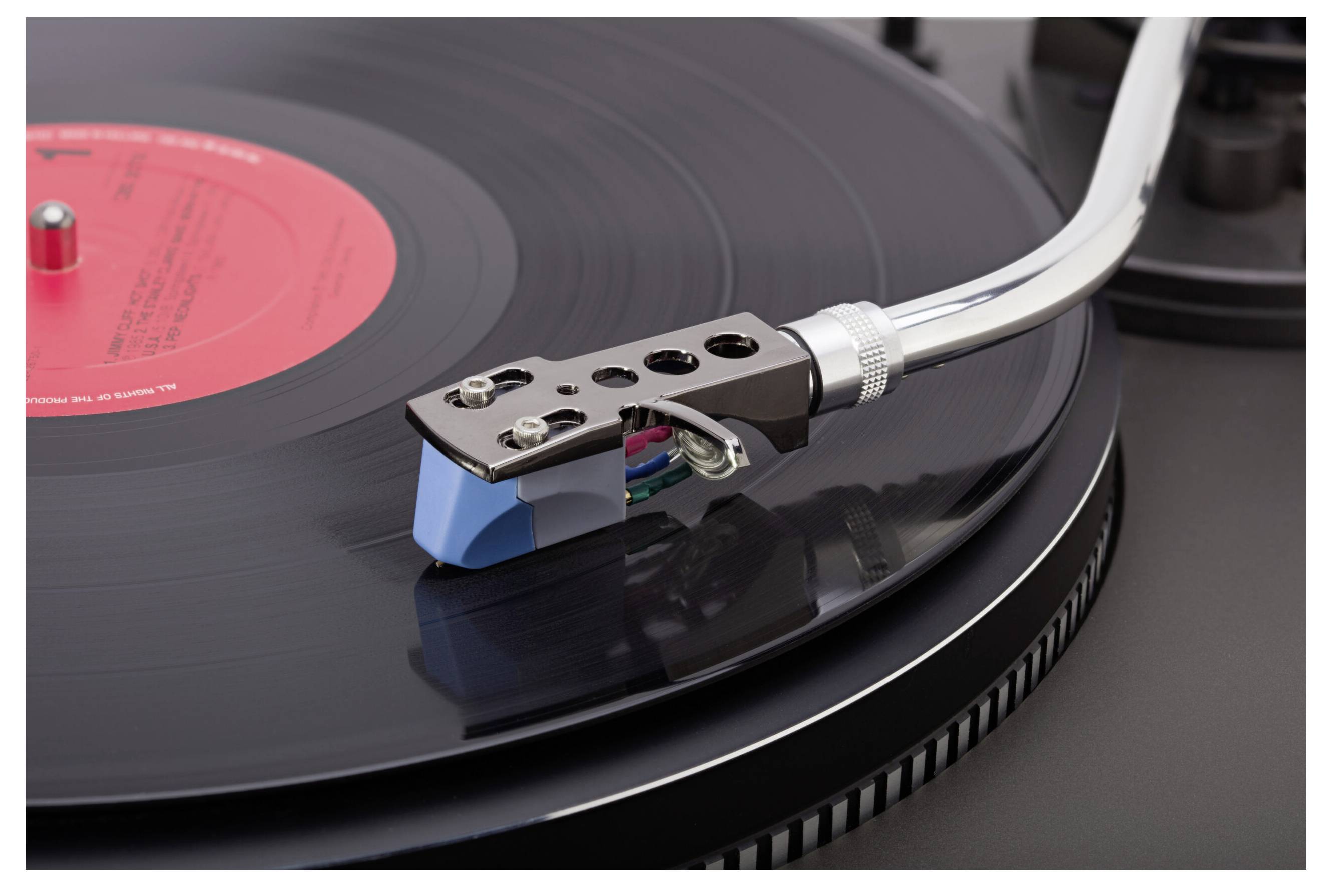 A close-up of a record player with a spinning black vinyl record under the needle and arm, highlighting the red label on the record.