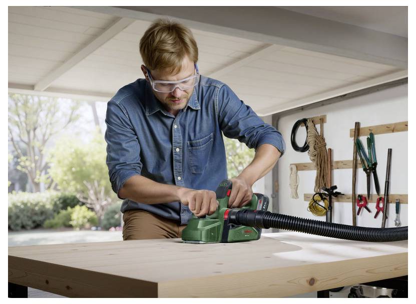 A person using an electric sander on a wooden table in a workshop, wearing safety goggles. Garden is visible through the window. Tools are hanging on the wall.