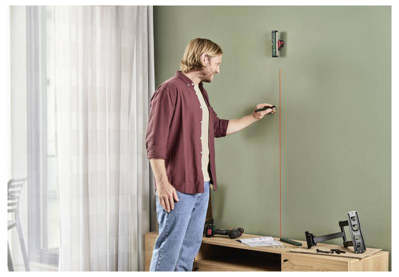 A person uses a laser level to mark a vertical line on a green wall. A drill and mounting equipment are on the table nearby.