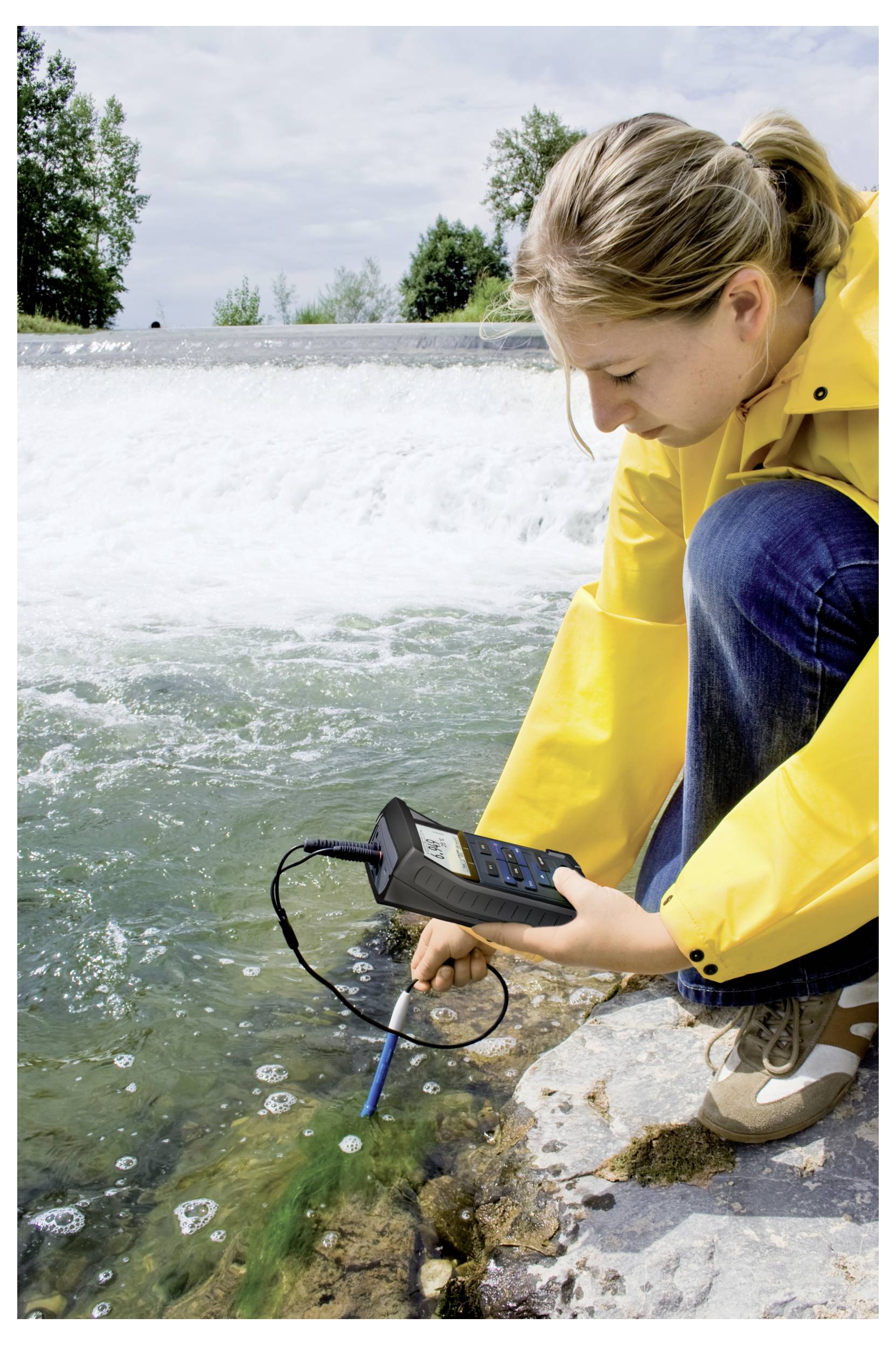 A person in a yellow raincoat uses a digital device to test water quality by a riverbank, indicating environmental data collection.