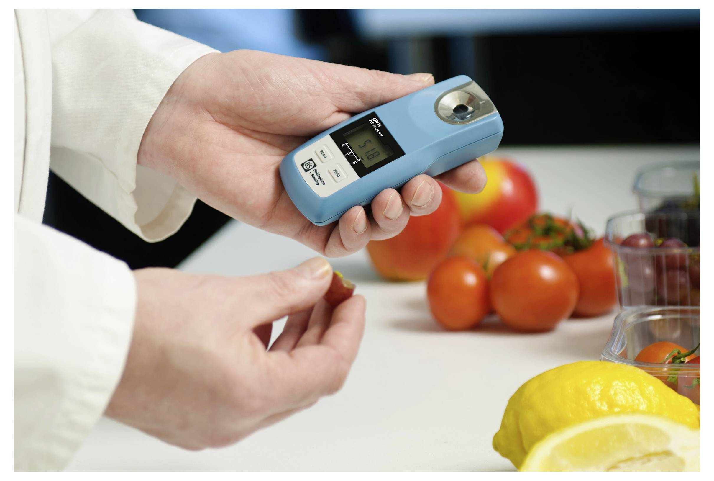 A person uses a digital refractometer to measure sugar levels in fruit. Tomatoes and lemons are on the table, indicating a laboratory setting.