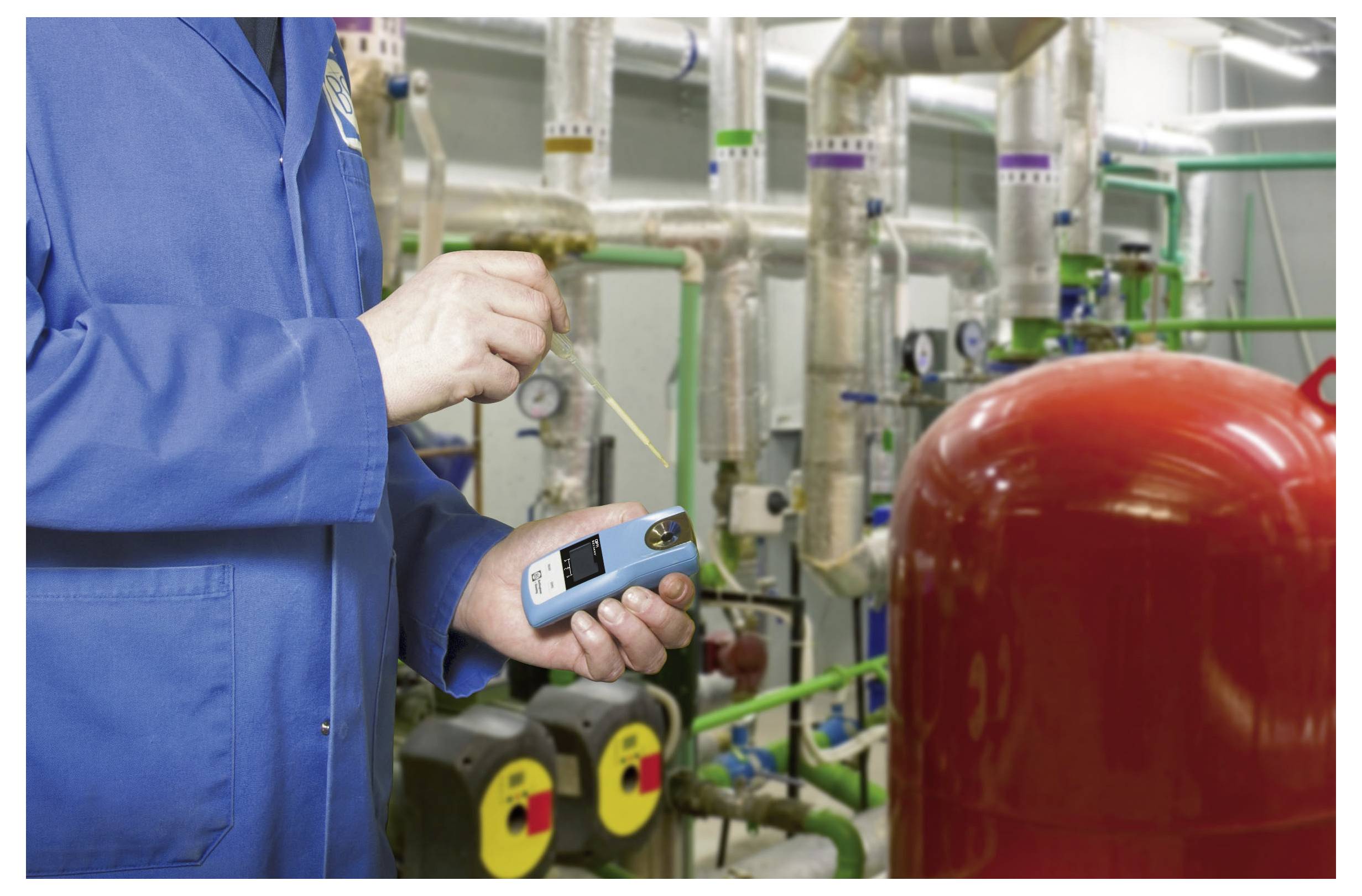 A person in a blue lab coat is using a handheld device to measure data in an industrial setting with pipes and machinery.
