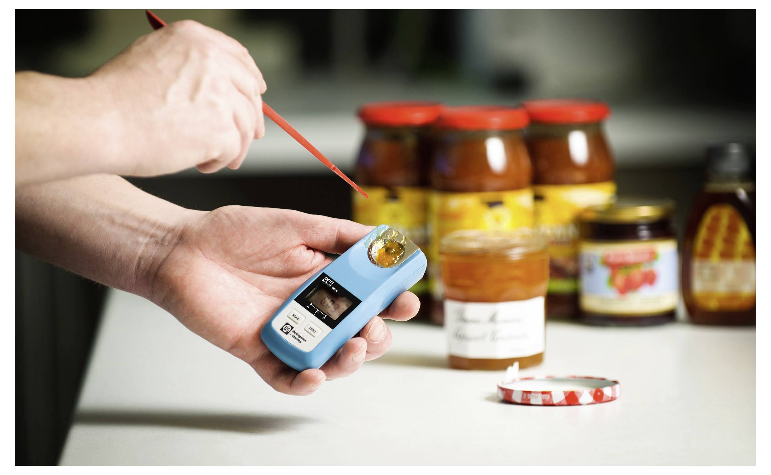 Hands testing honey with a refractometer in a kitchen, with jars of honey and preserves in the background.