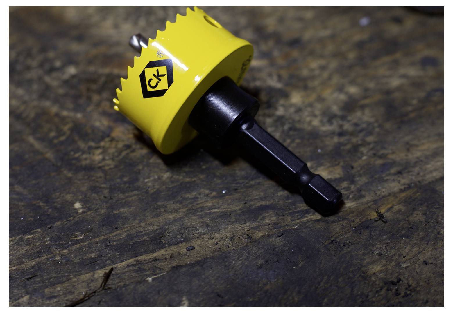 A yellow and black hole saw drill bit with a hex shank lies on a textured wooden surface, featuring a logo with initials.