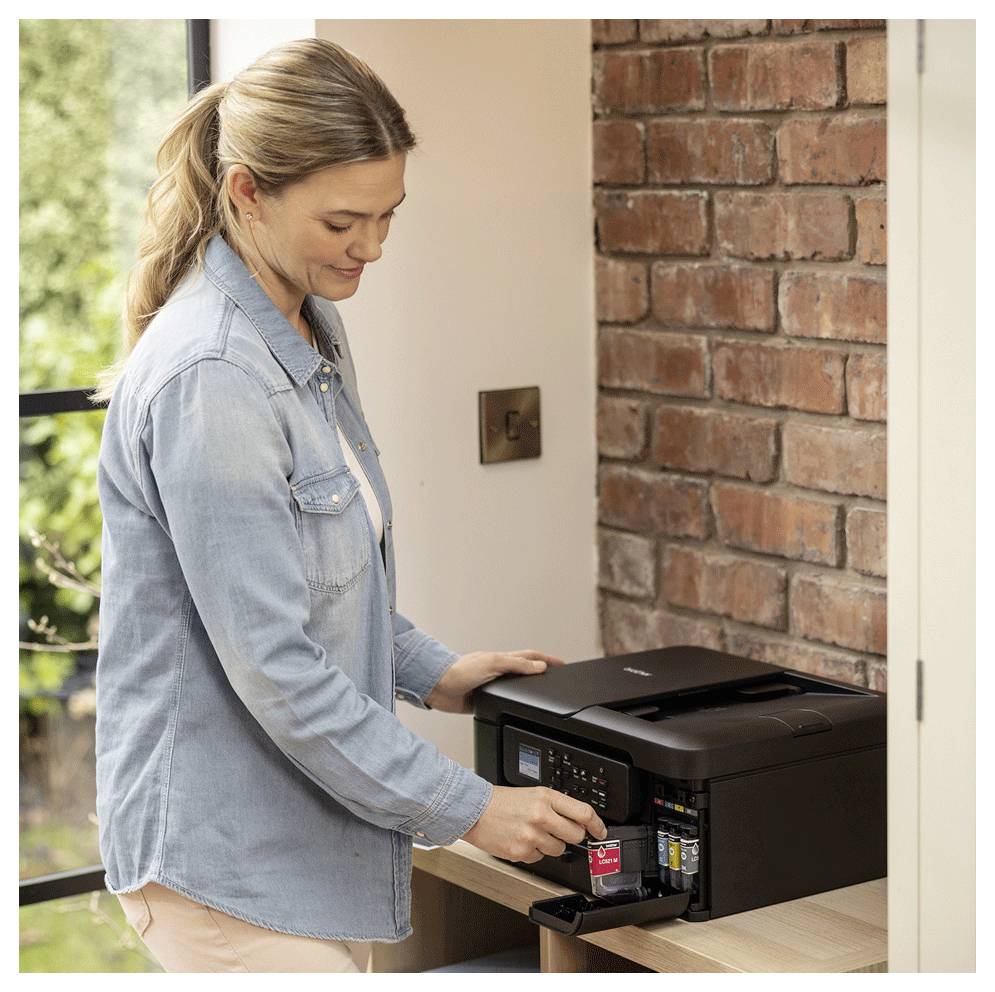 A woman inserts ink cartridges into a printer on a wooden shelf against a brick wall, indicating setup or maintenance of the device.