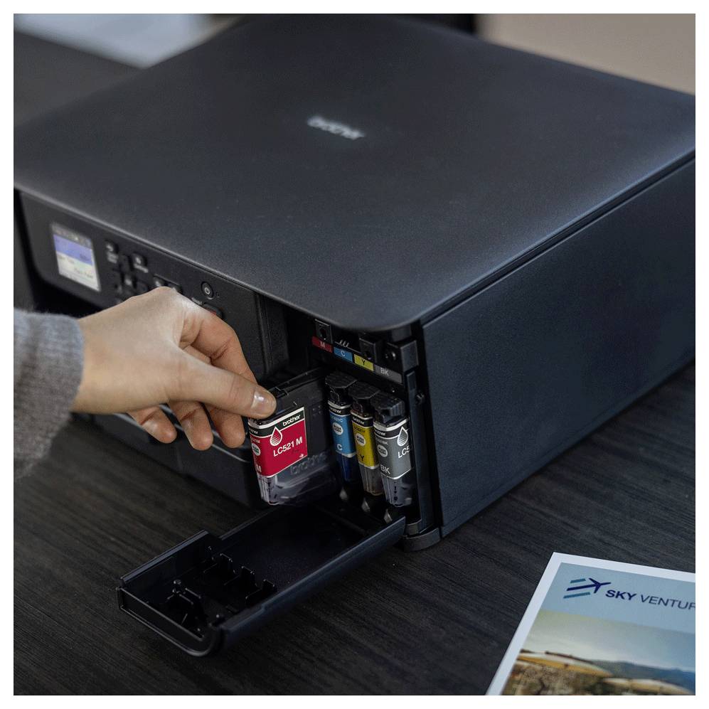 A person replaces a red ink cartridge in a printer, with a paper labeled 'Sky Venture' nearby, suggesting a printing setup.